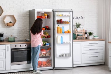 Genç bir kadın mutfaktaki yiyeceklerle dolu açık buzdolabına taze ananas koyuyor.