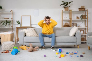 Stresli genç adam evde dağınık çocuk oyuncaklarıyla kanepede oturuyor.