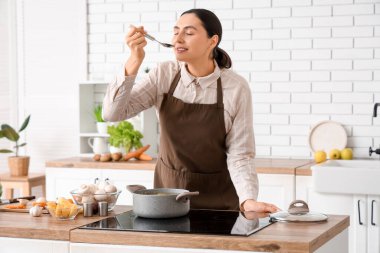 Genç bir kadın mutfakta çorba tadıyor.