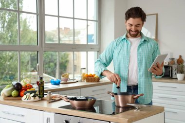 Tablet bilgisayarlı yakışıklı sakallı adam mutfakta ocakta yemek pişiriyor.