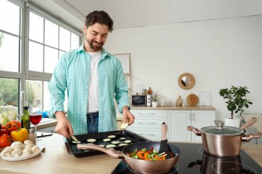 Yakışıklı sakallı adam mutfaktaki yemek tepsisine kabak dilimleri koyuyor.