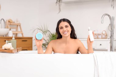 Güzel, genç ve mutlu bir kadın saç masörü ve bir şişe kozmetik ürünüyle evdeki küvette.