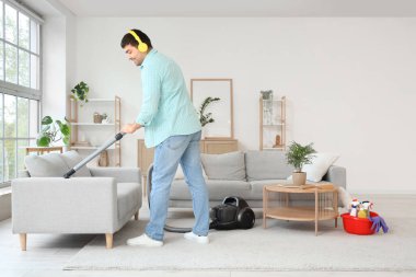 Genç adam kanepeyi süpürüyor ve odada müzik dinliyor.