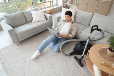 Evde dizüstü bilgisayarı ve elektrikli süpürgesi olan genç bir adam.