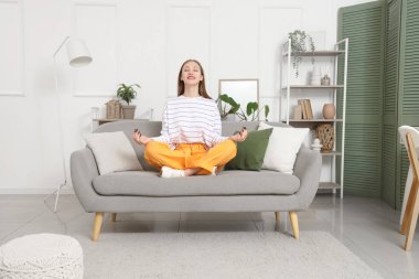 Genç bir kadın evdeki gri kanepede meditasyon yapıyor.