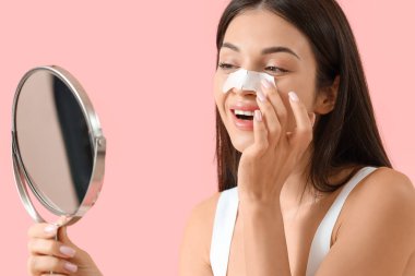 Burun bandı takmış genç bir kadın pembe arka planda aynaya bakıyor.