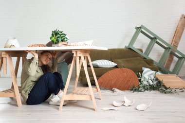 Depremden sonra dağınık oturma odasında masanın altında saklanan genç bir kadın.