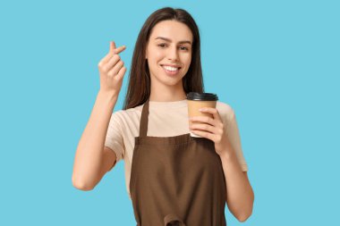 Kahveli genç bayan barista mavi arka planda kalp hareketi yapıyor.