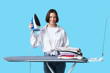 Mavi arka planda ütü masasında demir ve temiz giysiler yığını olan genç bir kadın.