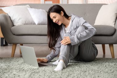 Gri pijamalı genç bir kadın evde dizüstü bilgisayarla çalışıyor.