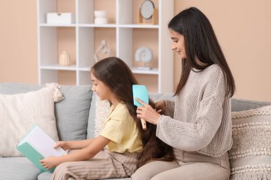 Genç anne, evdeki kanepede oturup kitap okuyan küçük kızının saçını tarıyor.