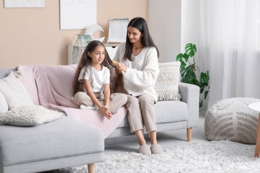 Genç anne, evdeki kanepede otururken küçük kızının saçını tarıyor.