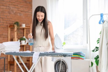 Sprey şişesi, ütüsü ve banyoda giyecek yığını olan genç ve güzel bir kadın.