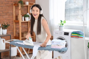 Kulaklıklı, evde giysilerini ütüleyen güzel, genç bir kadın.