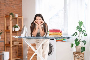 Güzel, genç ve mutlu bir kadın banyoda demir ve giysi yığınıyla.