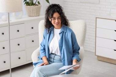 Şık mavi gömlekli, dergisi evdeki koltukta oturan güzel Afro-Amerikan kadın.