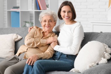 Gülümseyen hemşire ve yaşlı kadın evde kanepede sarılıyorlar.