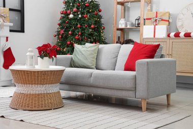 Oturma odasının içinde kanepe, köknar ağacı ve masa üzerinde Noel çiçeği süsleri var.