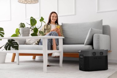 Evinde taşınabilir elektrik santrali olan koltukta cep telefonu kullanan genç bir kadın.