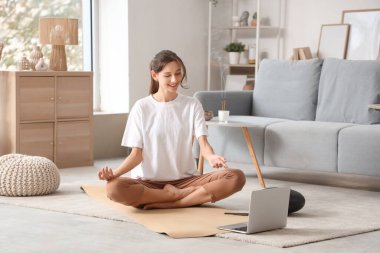 Evde yoga minderi üzerinde meditasyon yapan genç bir kadın.