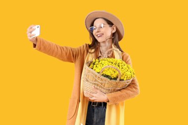 Kasımpatı çiçekli gözlüklü şık genç bir kadın sarı arka planda selfie çekiyor.