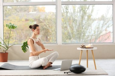 Bilgisayarlı genç bir kadın pencerenin yanında meditasyon yapıyor.