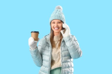 Kışın giyinmiş, kahve fincanı olan genç bir kadın mavi arka planda cep telefonuyla konuşuyor.