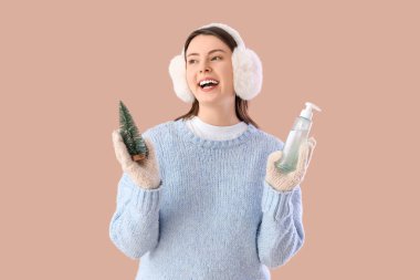 Cilt bakımı ürünü ve bej arka planda oyuncak Noel ağacıyla kışın giyinmiş genç bir kadın.