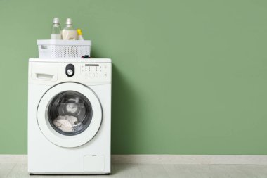 Çamaşır makinesi. Yeşil duvarın yanında çamaşır deterjanı şişeleri var.