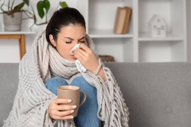 Evdeki kanepede hapşıran peçete ve bir fincan çayla hasta genç bir kadın.