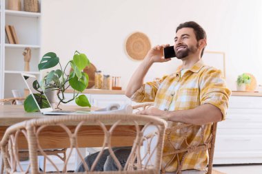 Mutfaktaki masada laptopuyla cep telefonuyla konuşan yakışıklı bir adam.