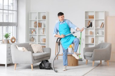 Önlüklü genç adam elektrikli süpürgeyle evde eğleniyor.