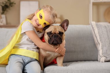 Evdeki kanepede oturan süper kahraman kostümlü Fransız bulldog 'u olan şirin mutlu küçük kız.