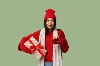 Güzel, genç ve mutlu bir Asyalı kadın kışlık giysiler içinde sıcak şarap ve yeşil arka planda hediye kutusuyla.