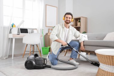 Kulaklıklı yakışıklı adam evdeki kanepenin altındaki süpürgeyi temizliyor.