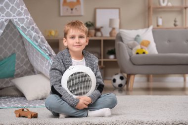 Evde yerde elektrikli vantilatör ısıtıcısı olan şirin bir çocuk oturuyor.