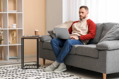 Genç serbest çalışan oturma odasındaki kanepede dizüstü bilgisayar kullanıyor.