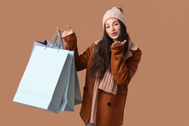 Güzel, genç bir kadın, sıcak kıyafetler içinde, alışveriş torbalarıyla kahverengi arka planda öpüşüyor.