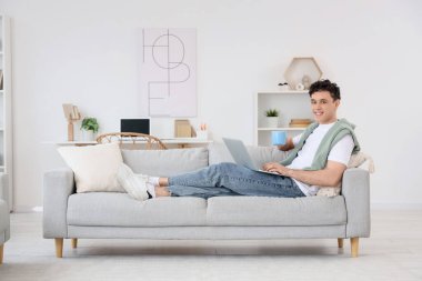 Evdeki kanepede dizüstü bilgisayarla çalışan genç adam.