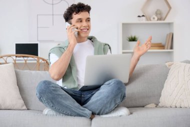 Bilgisayarlı genç adam evdeki koltukta cep telefonuyla konuşuyor.