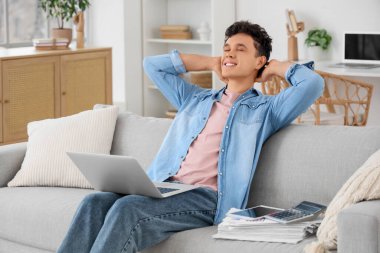 Dizüstü bilgisayarı olan genç adam evdeki kanepede dinleniyor.