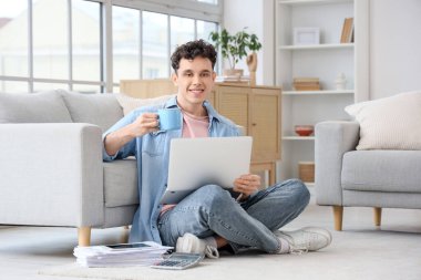 Genç adam evde dizüstü bilgisayar ve bir fincan kahveyle çalışıyor.