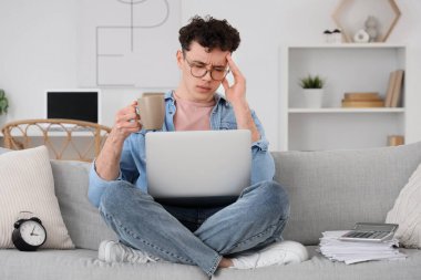 Evdeki kanepede oturan bir fincan kahve ve dizüstü bilgisayarlı stresli genç adam.