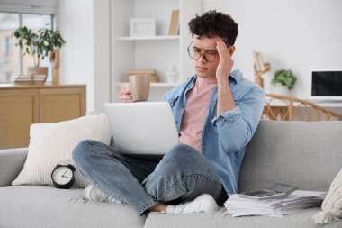 Evdeki kanepede oturan bir fincan kahve ve dizüstü bilgisayarlı stresli genç adam.