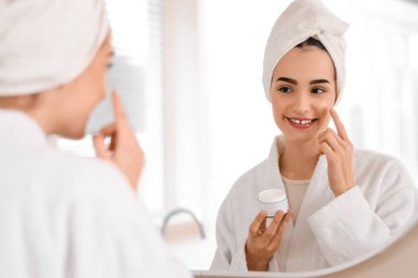 Banyoda duştan sonra aynanın yanında krem süren genç bir kadın.