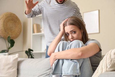 Kanepede oturan genç bir kadın ve evde oturan kocası. Aile içi şiddet kavramı