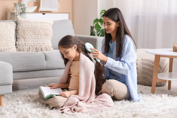 Genç anne evde oturmuş kitap okuyan küçük kızının saçlarını tarıyor.