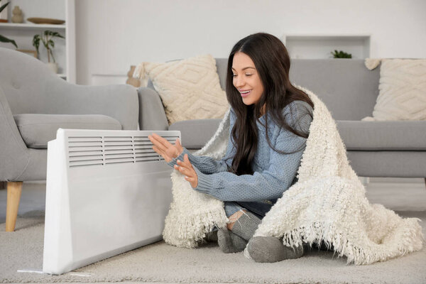 Frozen young woman with plaid warming hands near electric convector heater at home