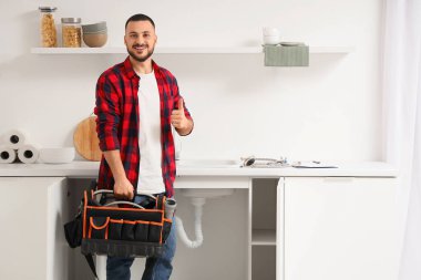 Alet çantalı erkek tesisatçı mutfaktaki lavabonun yanında baş parmağını kaldırıyor.