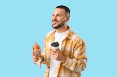 Elinde bir fincan kahve ve mavi arka planda kruvasan olan genç adam.